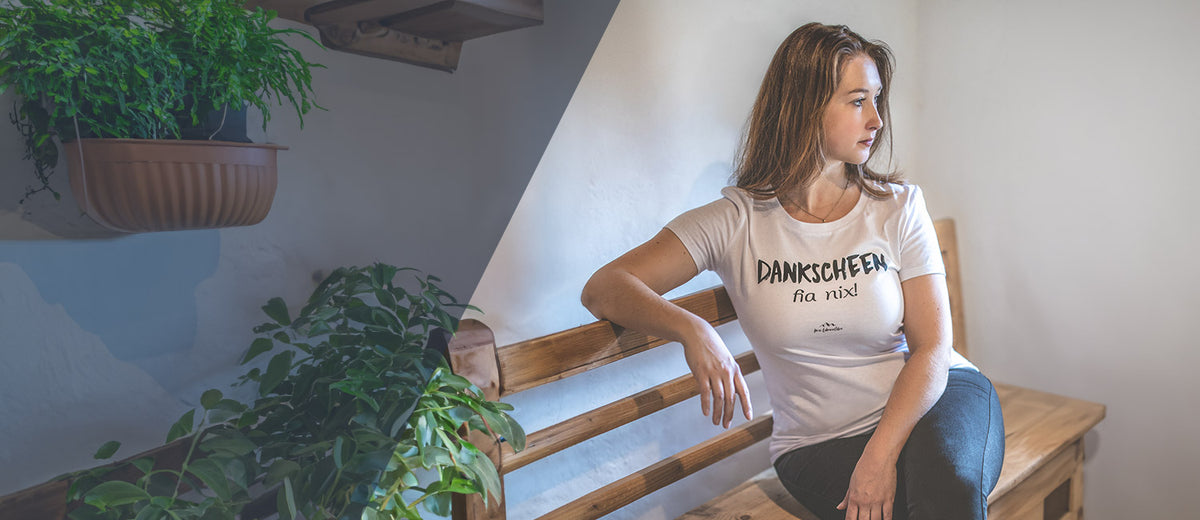Junge Frau trägt weißes Trachten-Shirt mit bayerischem Spruch Dankscheen fia nix auf Holzbank sitzend.