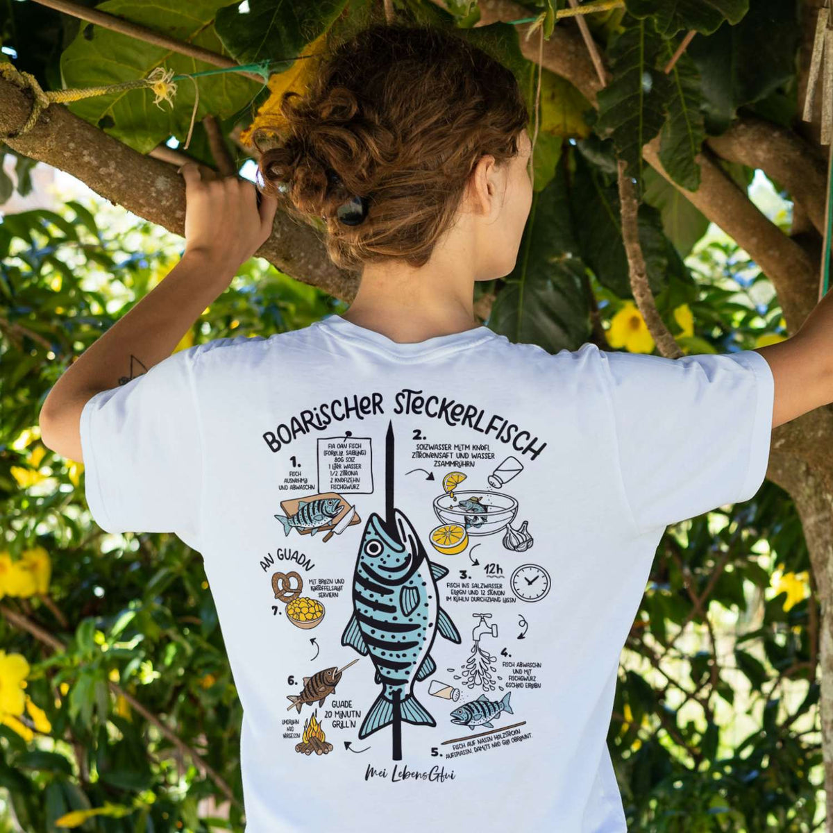 Rückenansicht eines Damen T-Shirts "Boarischer Steckerlfisch" mit Rezeptanleitung unter Bäumen.