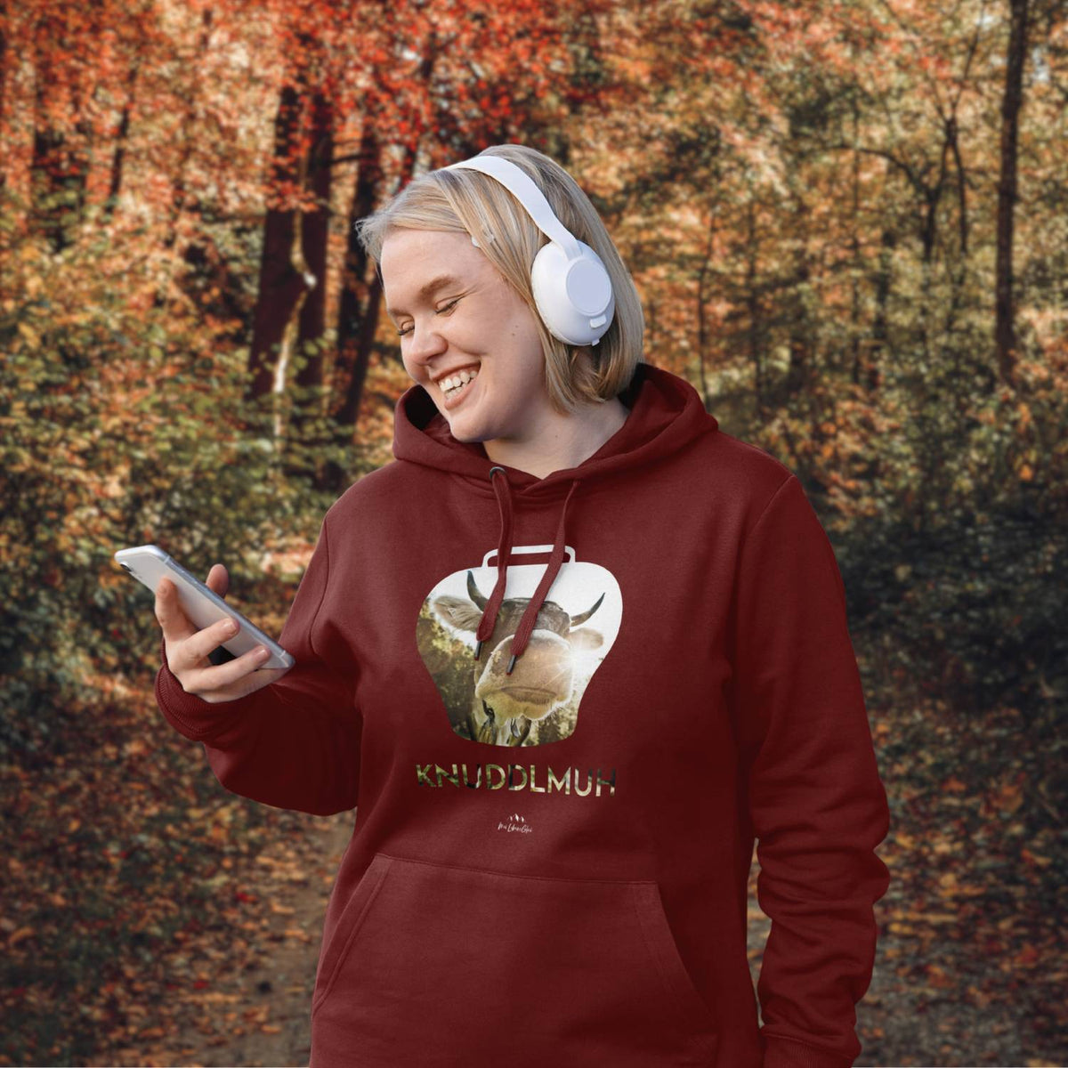Lächelnde Frau mit weißem Kopfhörer und rotem Hoodie Knuddlmuh mit Kuhmotiv im herbstlichen Wald.