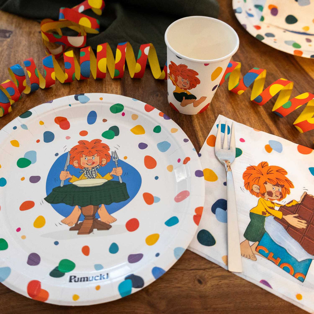 Pumuckl Partyset auf dem Geburtstagstisch: Pappteller mit Pumuckl beim Essen, Pappbecher und Serviette mit Schokoladen-Motiv, dazu bunte Luftschlangen