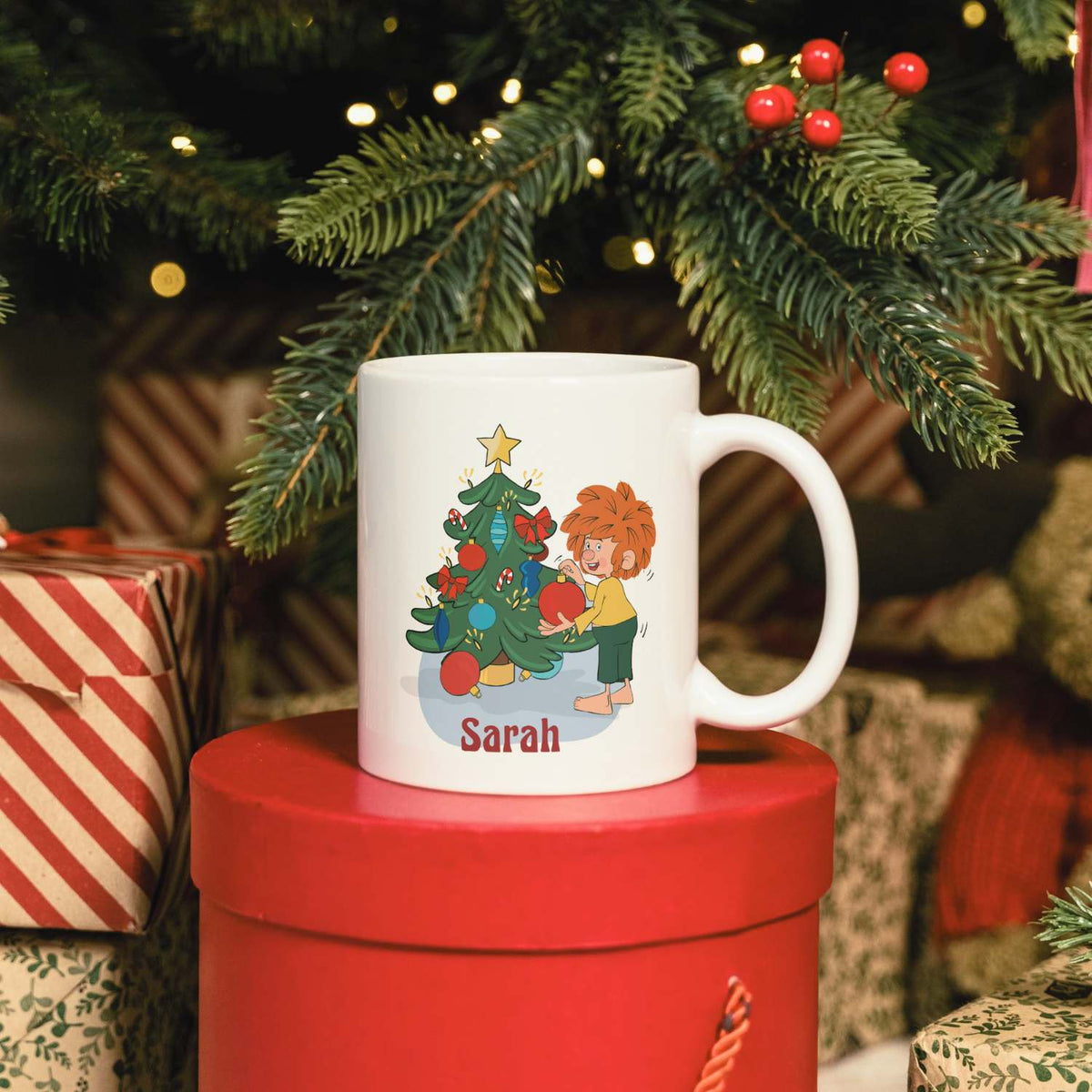 Weiße ®Pumuckl Tasse Weihnachten personalisierbar mit Pumuckl am Christbaum, steht auf roten Geschenkboxen.