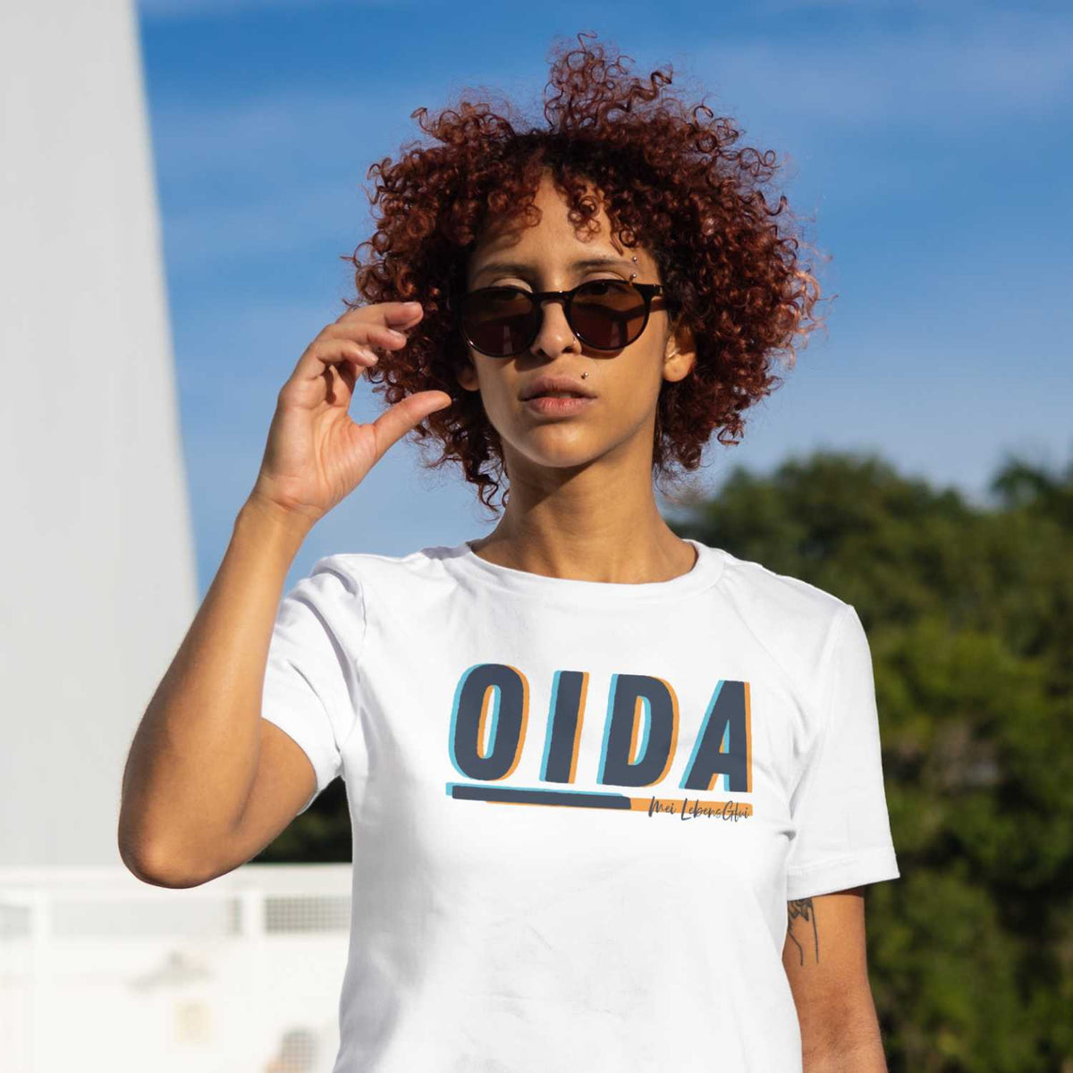 Junge Frau mit Locken und Sonnenbrille trägt weißes Damen T-Shirt Oida mit blau-orangefarbenem Schriftzug.
