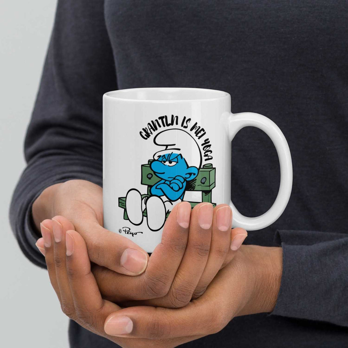 Weiße ®Die Schlümpfe Tasse "Grantln is mei Yoga" mit mürrischem Schlumpf in den Händen einer Person.