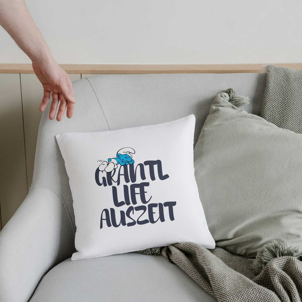 Weißes ®Die Schlümpfe Grantl Life Auszeit Kuschelkissen auf grauem Sessel mit Decke, eine Hand berührt den Stuhl.