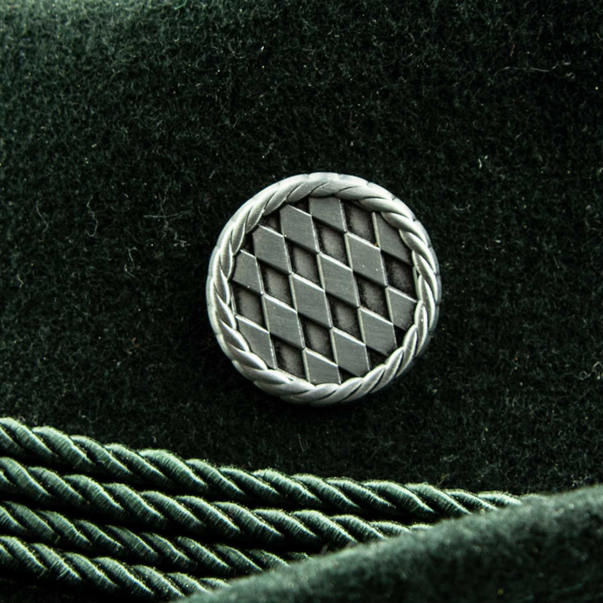 Pin Bayrische Rauten, rund, mit Silberrand und Rautenmuster, auf dunkelgrünem Filzstoff.