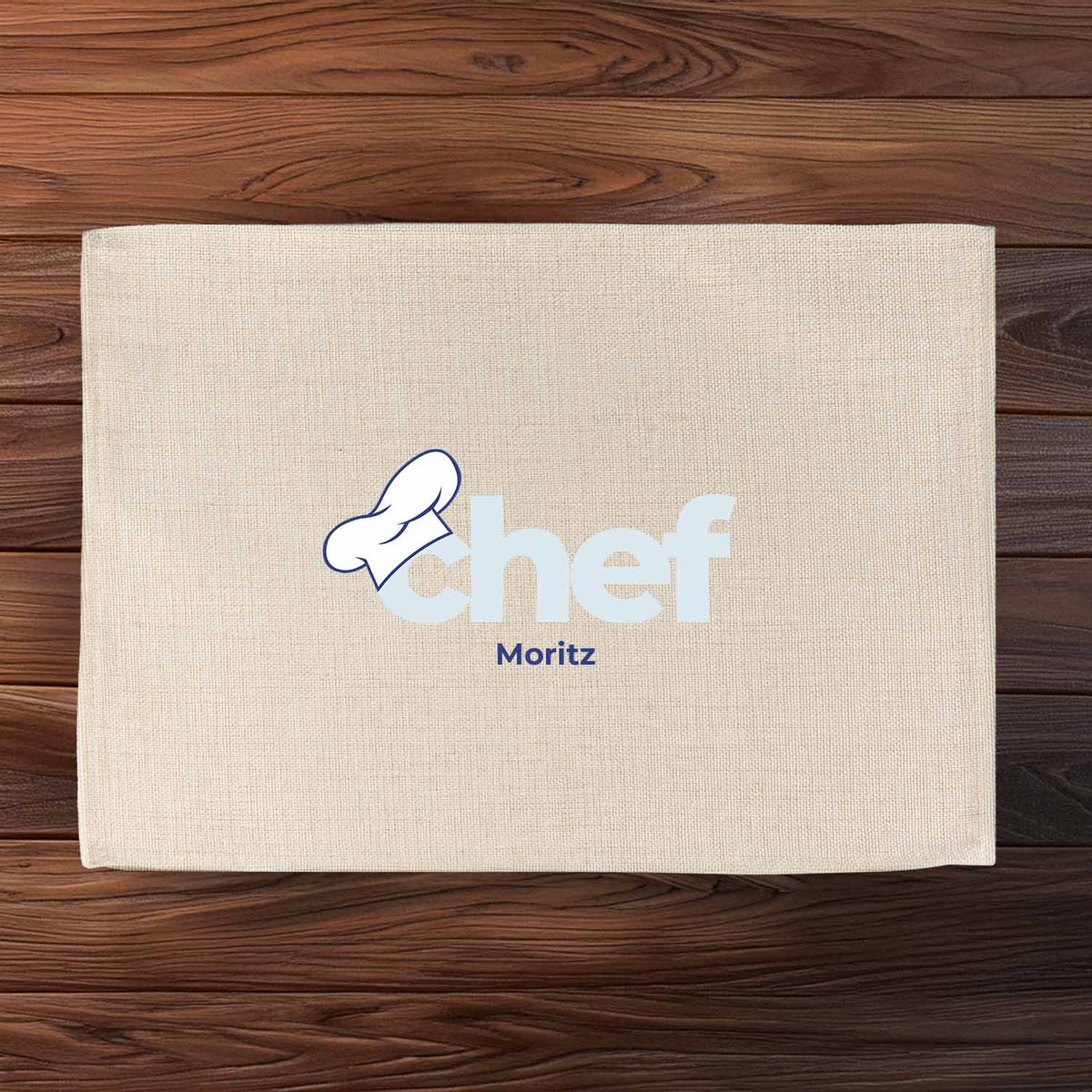 Beiges Tischset "Chef" mit Kochmütze und dem Personalisierungsbeispiel "Moritz" auf rustikaler Holzoberfläche.