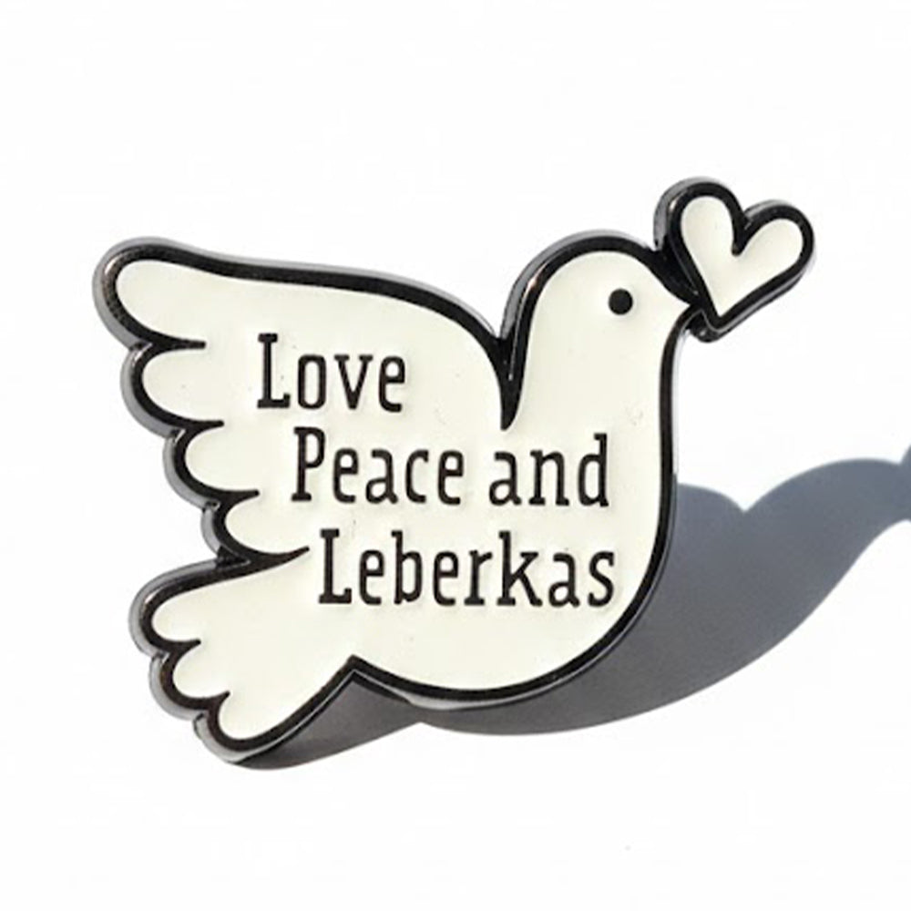 Pin „Love, Peace and Leberkas“: weiße Emaille-Friedenstaube mit Herz im Schnabel und schwarzem Schriftzug