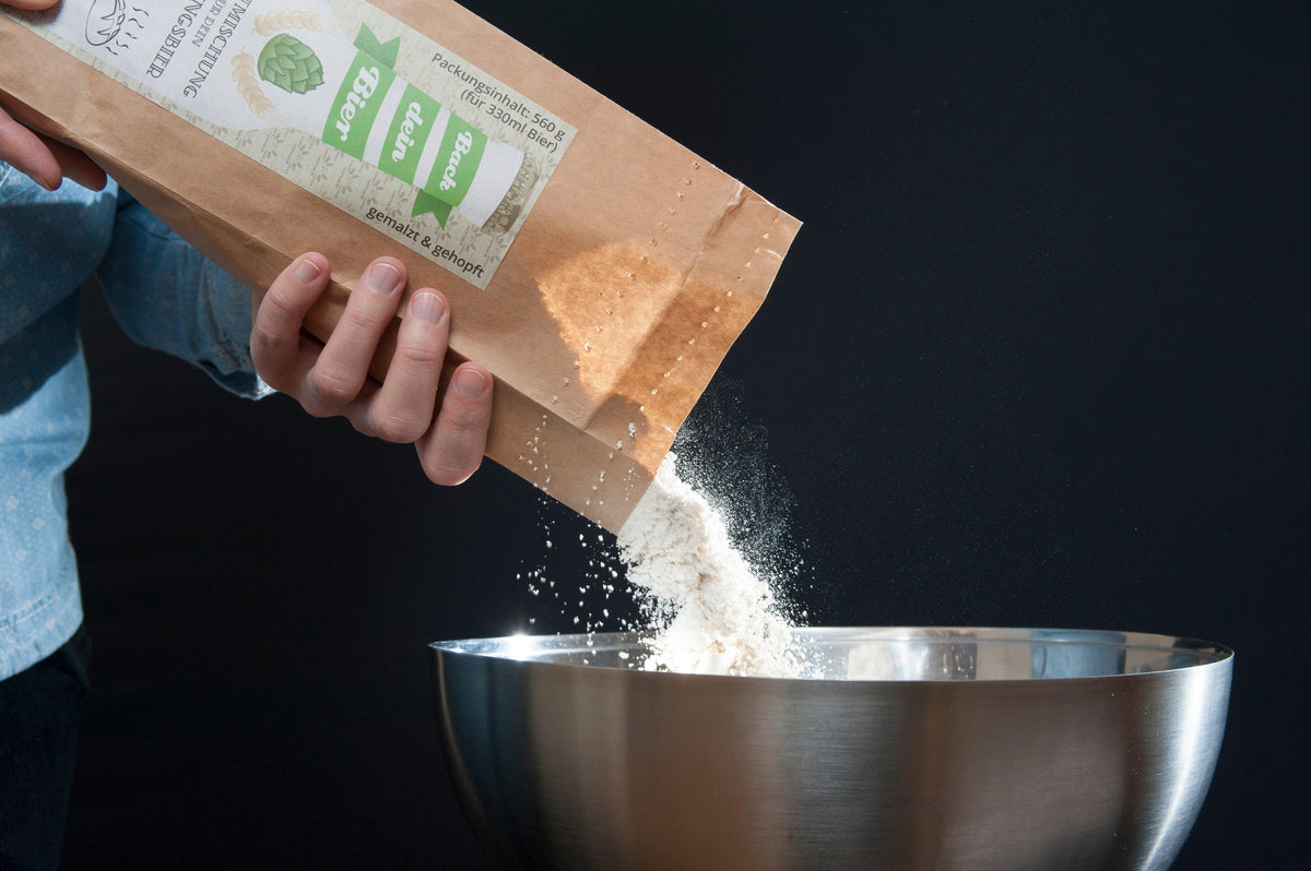 Person schüttet "Back dei Bier" Brotmischung aus Papiertüte in Edelstahlschüssel, fertig zum Backen.