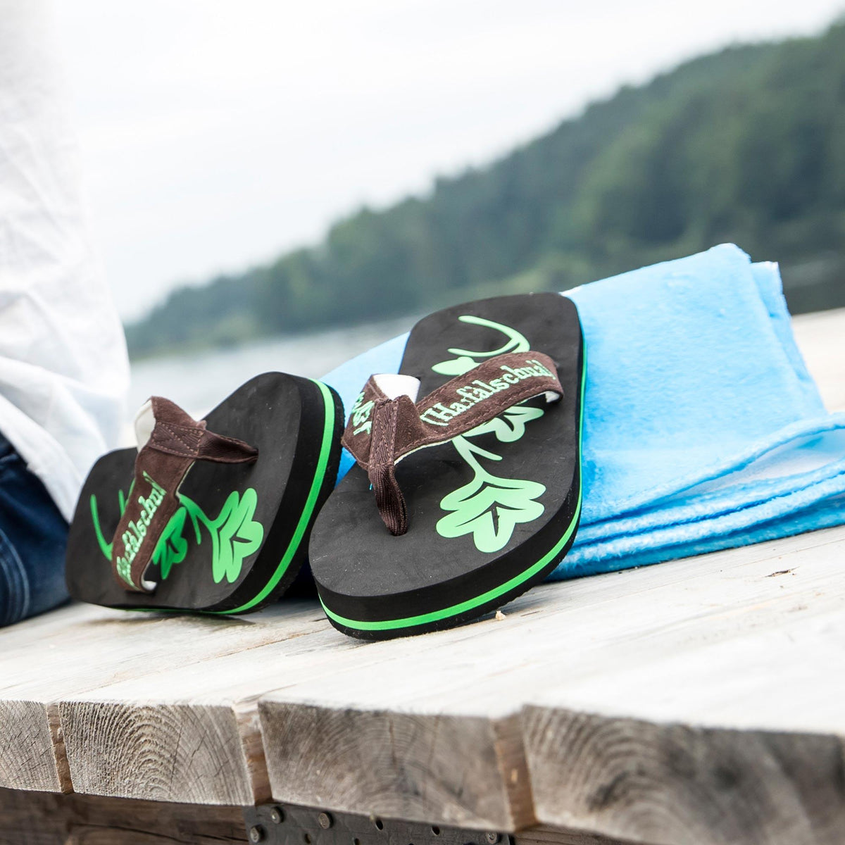 Badesandale Haferlschuh: Trachten Flip Flops für Herren mit Eichenlaub-Fußbett neben blauem Handtuch auf einem Holzsteg am See