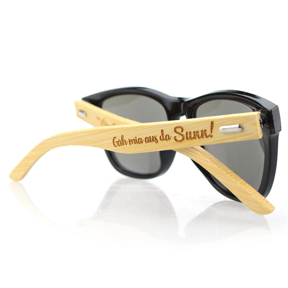Moderne Sonnenbrille mit schwarzem Gestell und dunklen Gläsern, Bügel aus hellem Bambusholz mit Gravur "Geh mia aus da Sunn!