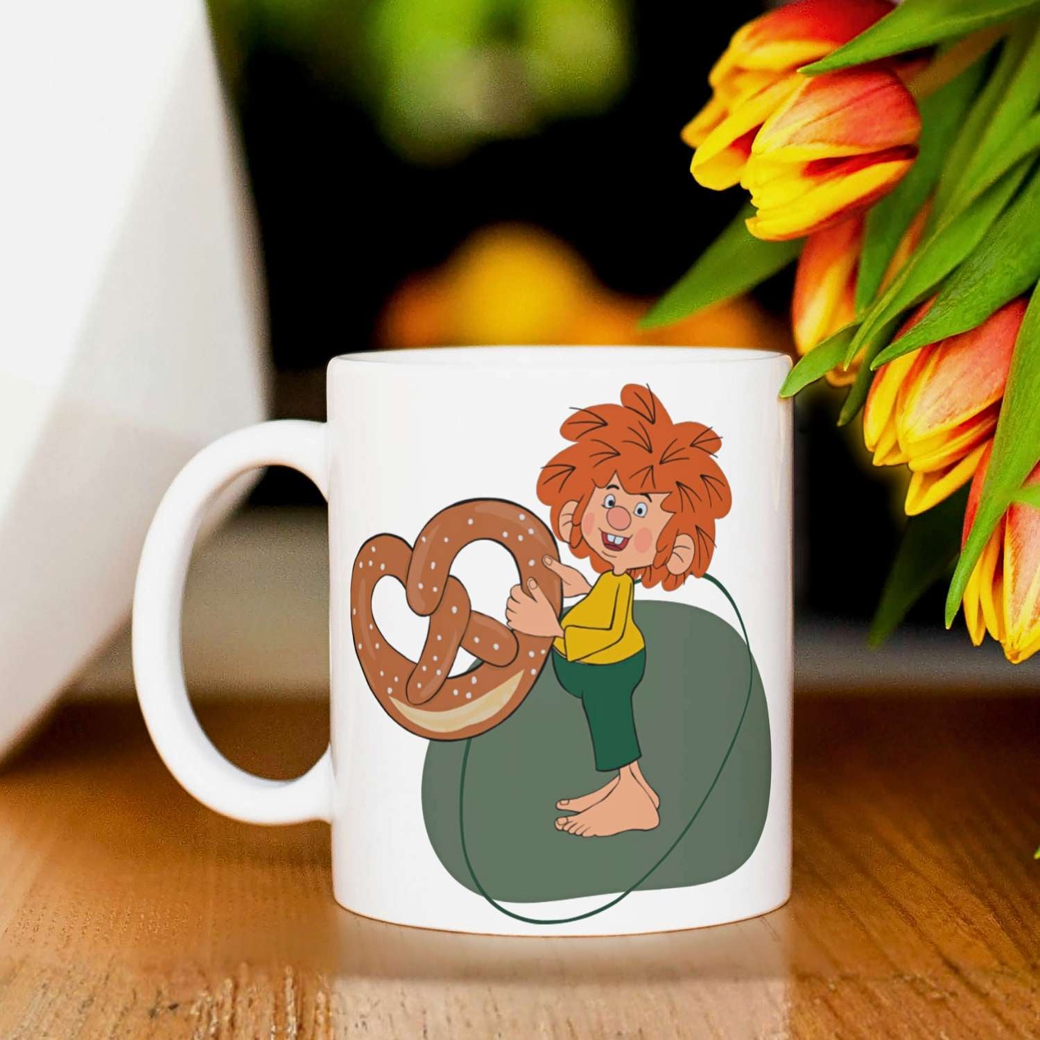 Weiße Pumuckl Tasse "Breze" mit rothaarigem Kobold, der eine Brezel hält, auf Holztisch.