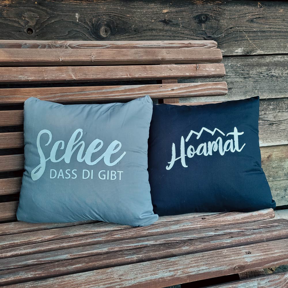 Zwei dekorative Kissen mit Aufdruck "Schee dass di gibt" (grau) und "Hoamat" (blau) auf Holzbank.
