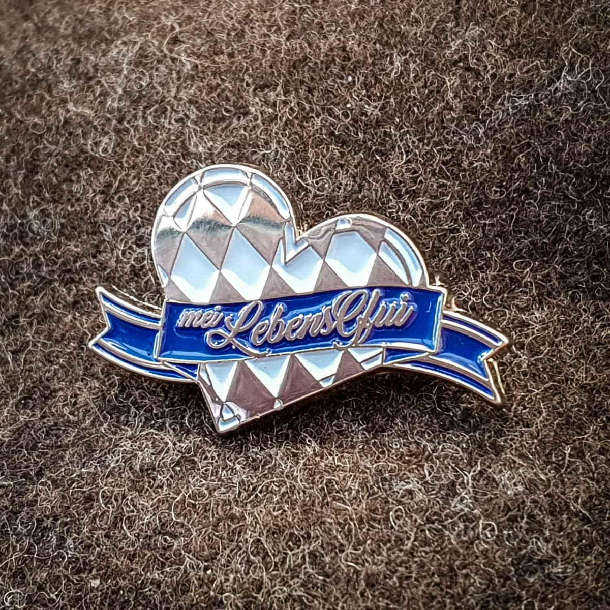 Ovaler Pin mit schwarzem Schriftzug auf silberfarbenem Hintergrund, aus Nickel, ca. 3×2 cm, mit Sicherheitsnadel.