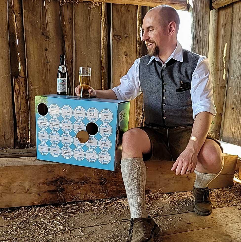 Mann in Tracht trinkt Bier neben der hellblauen Bierbox „Bayerischer Biergarten“ auf einem Holzbalken