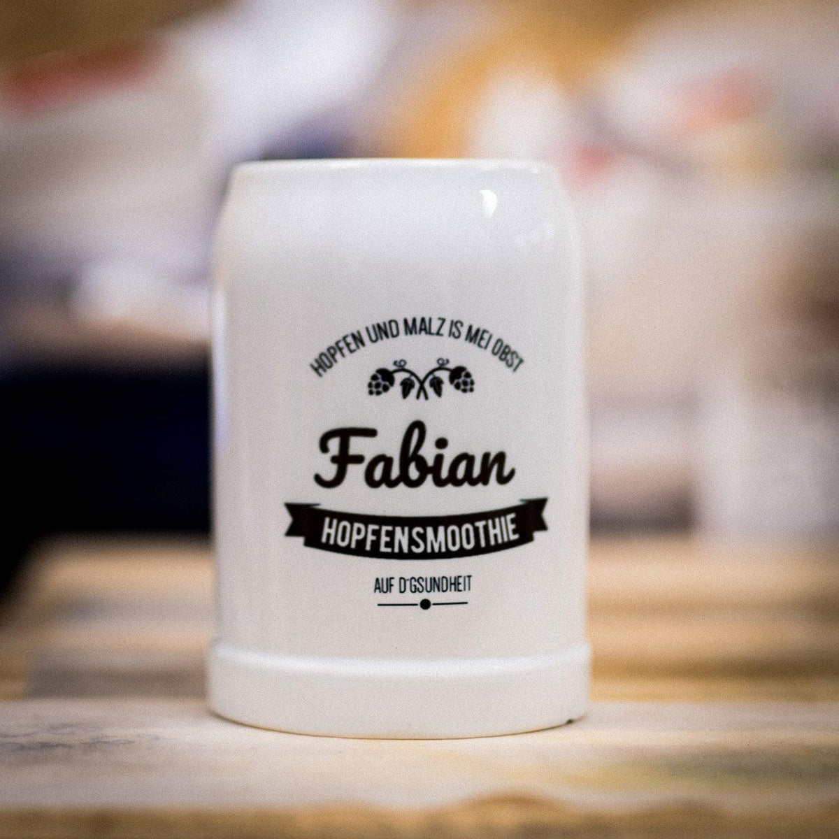 Weißer Bierkrug aus Porzellan mit schwarzem Aufdruck "Hopfensmoothie Fabian" und bayerischem Spruch auf hellem Holztisch.