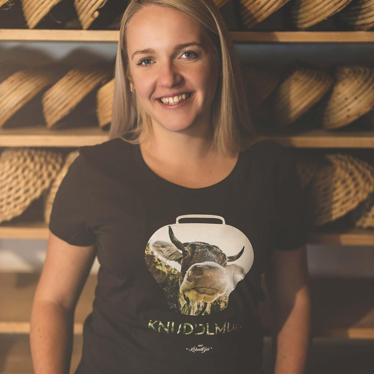 Lächelnde Frau in schwarzem Damen T-Shirt Knuddlmuh mit Kuh-Ausschnitt, umgeben von Weidenkörben.