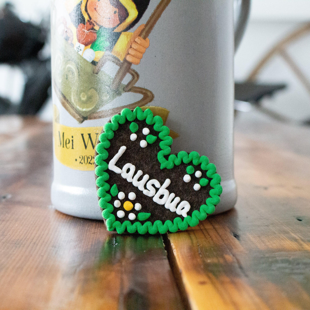 Braunes Filzherz-Anstecker "Lausbua" mit grüner Umrandung und weißen Punkten vor Bierkrug auf Holztisch.