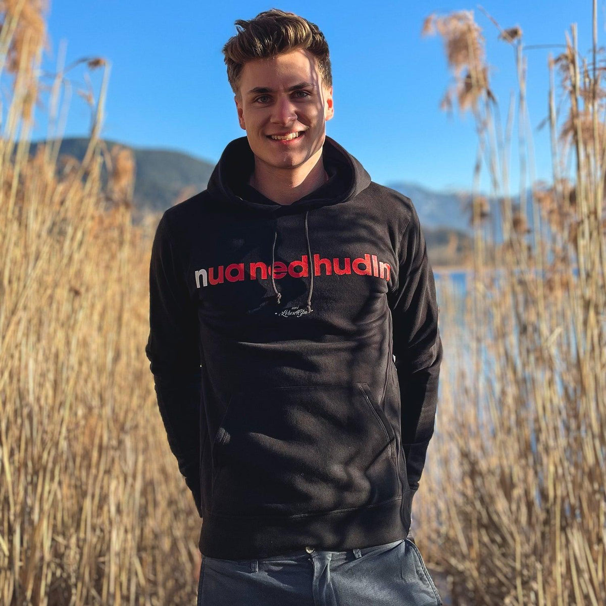 Lächelnder Mann im schwarzen Herren Hoodie Nua ned hudln mit rotem Schriftzug, stehend im sonnigen Schilf.