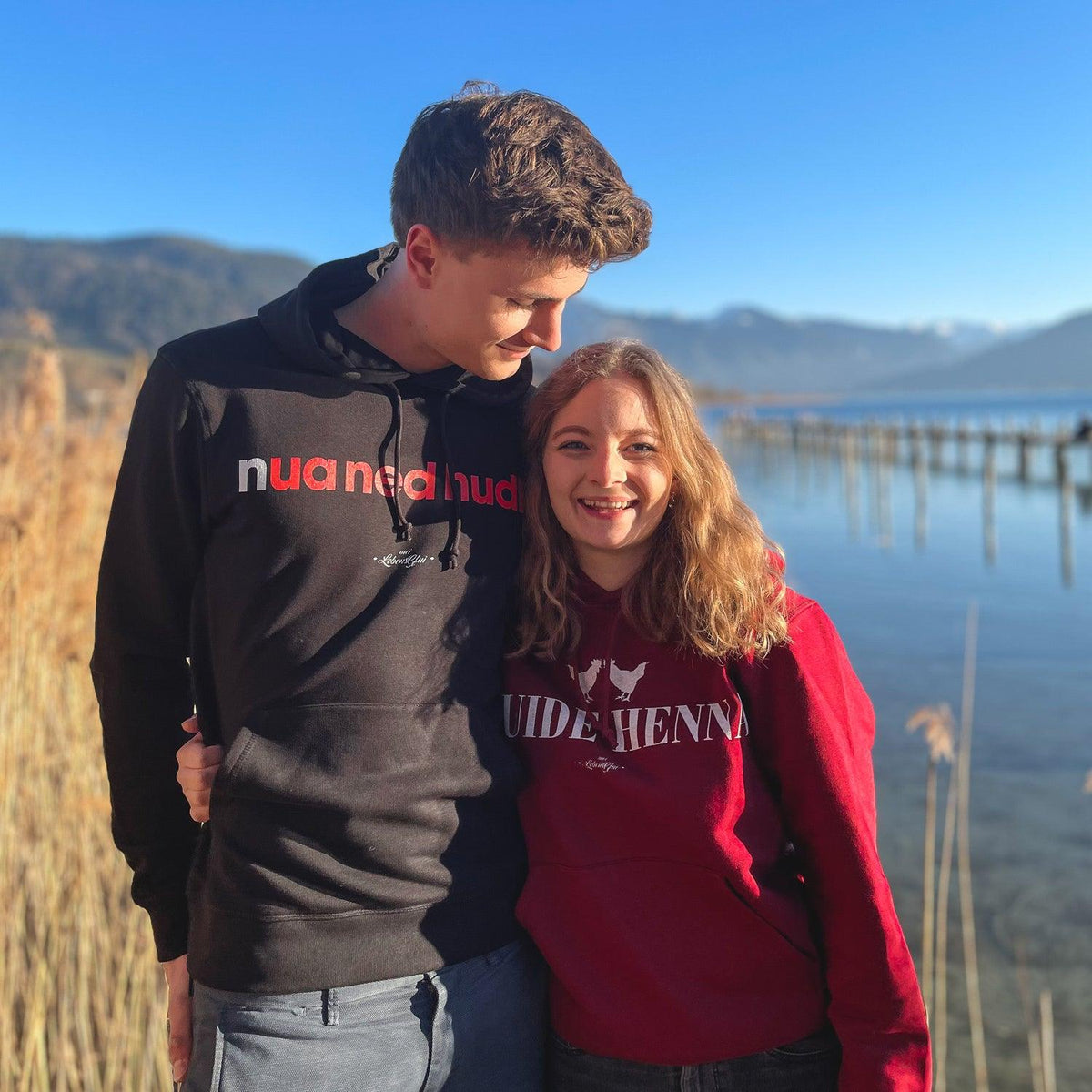 Junges Paar am See, er trägt einen schwarzen Herren Hoodie "Nua ned hudln" mit rotem Schriftzug, sie einen roten Hoodie.
