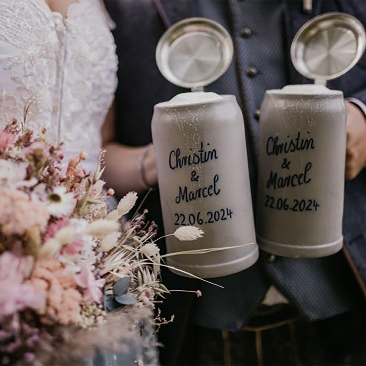 Paar hält zwei gravierte PREMIUM Bierkrug Steinkrüge mit Namen und Hochzeitsdatum, neben Blumen.
