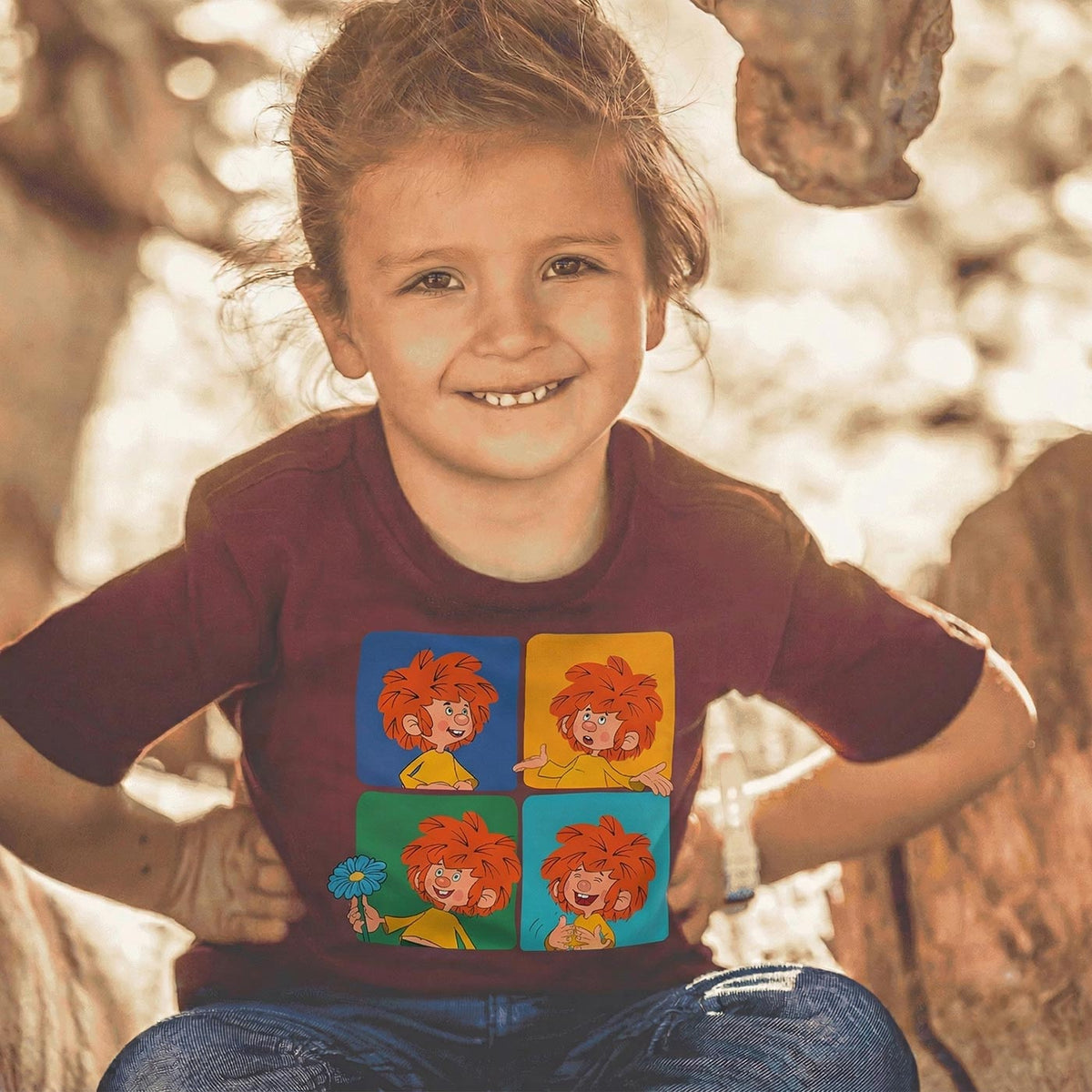 Lachendes Kind trägt bordeauxrotes Pumuckl T-Shirt mit vier Pumuckl-Motiven in bunten Quadraten