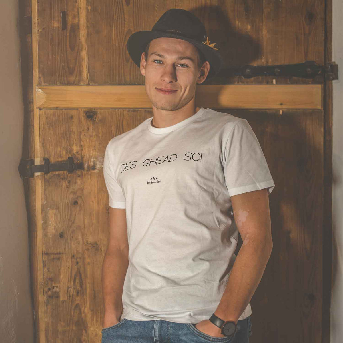 Junger Mann im schwarzen Trachtenhut und weißem T-Shirt mit Aufschrift vor Holztür lächelnd