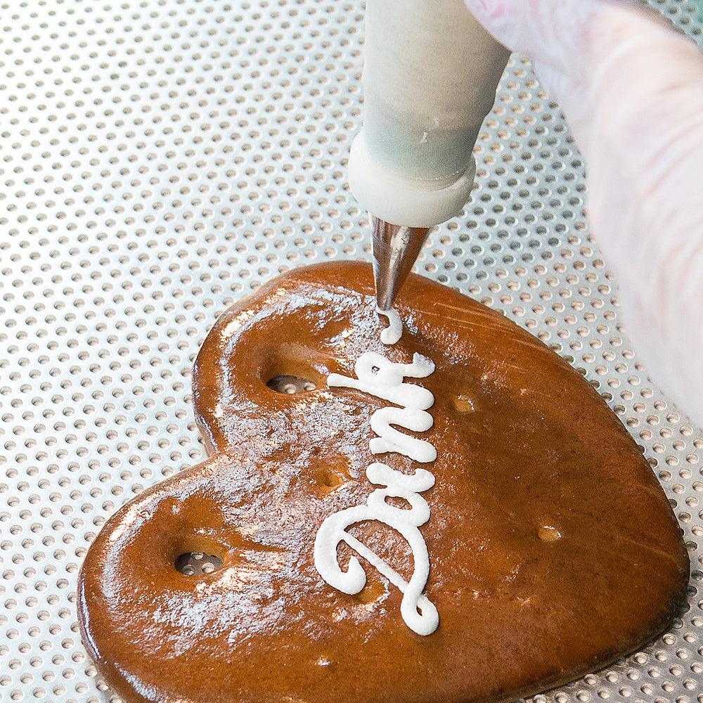 Konditorhand verziert herzförmiges Lebkuchenherz mit weißer Zuckerguss-Schrift Dank von Spritzbeutel.