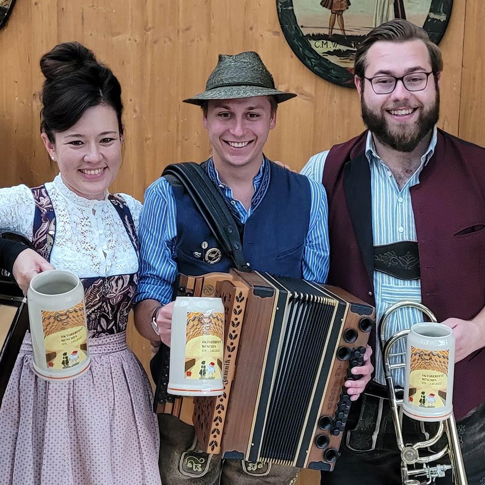 Drei fröhliche Musiker in Tracht halten den Offiziellen Oktoberfestkrug 2022 mit thematischen Motiven.