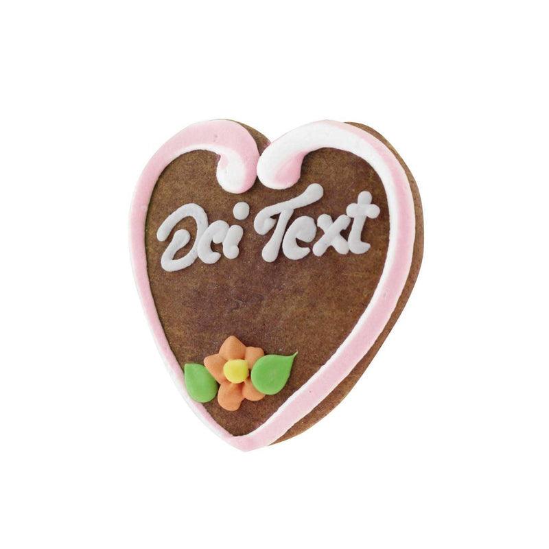 Braunes Lebkuchenherz mit weißer und rosa Zuckergussumrandung, bedruckt mit dem Wort "Dei Text" und orangefarbener Blume.