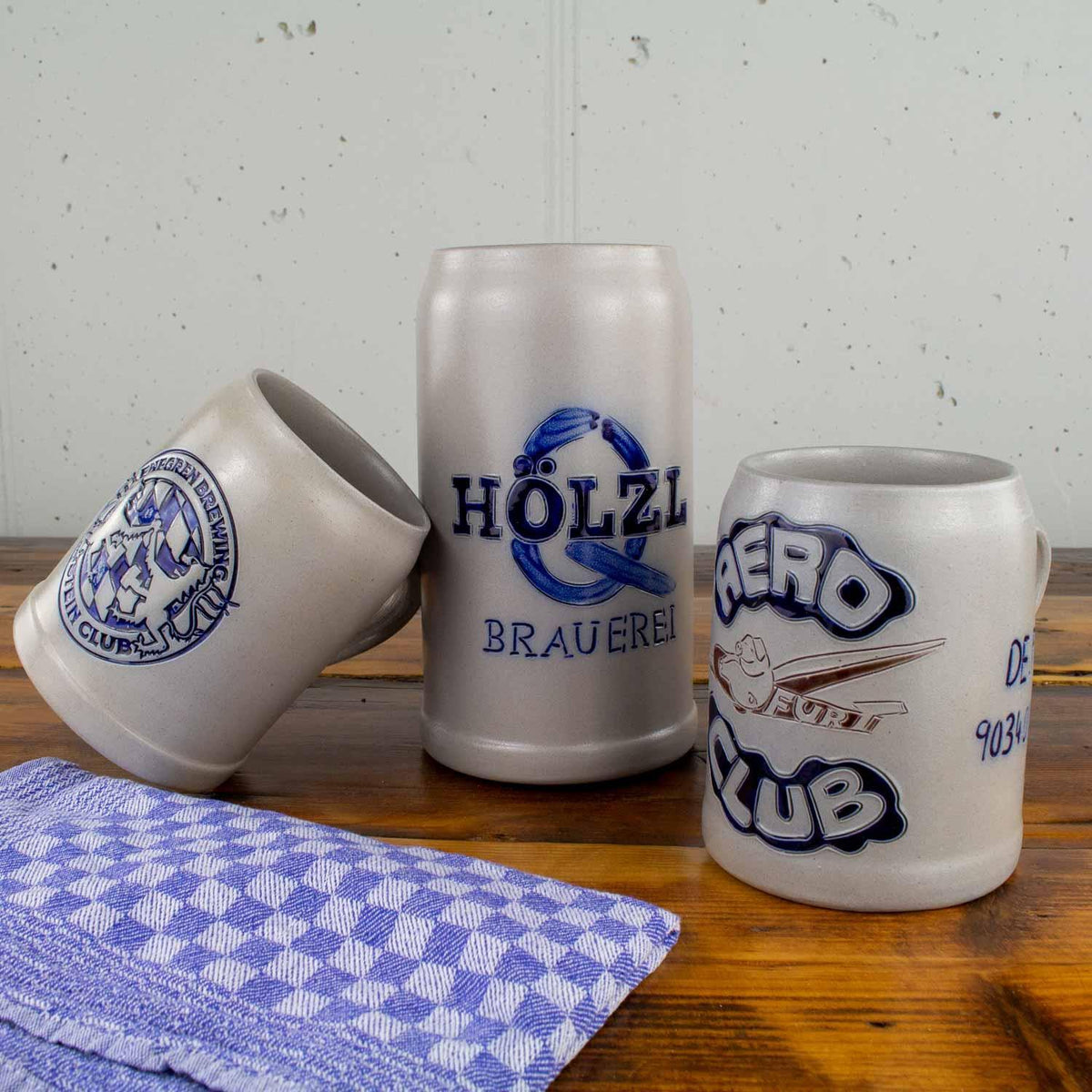 Verschiedene Keramik-Bierkrüge und Steinzeug-Bierseidel mit blauen Logos auf Holztisch und kariertem Handtuch.