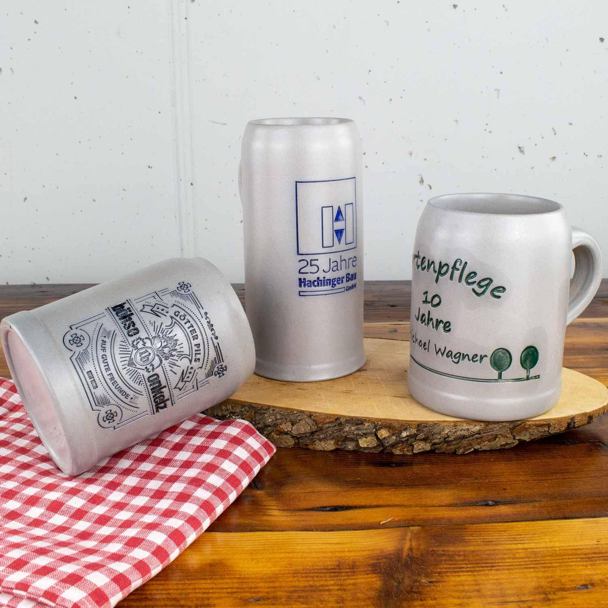 Verschiedene personalisierte Steinkrüge mit Logo und bayerischem Bierkrug-Design auf Holzbrett und rot-weiß karierter Serviette.