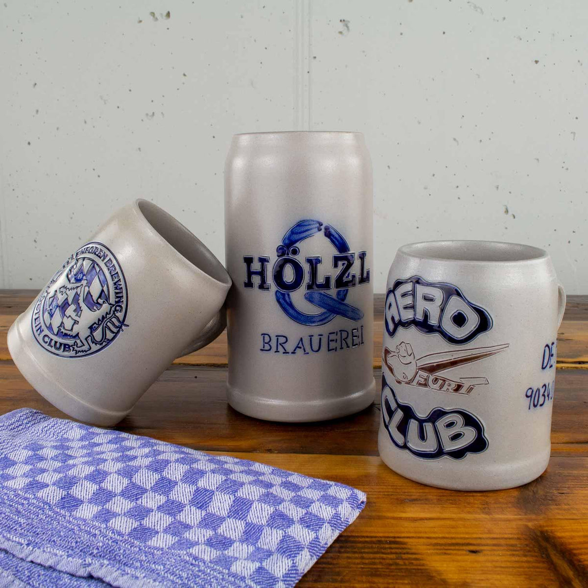 Drei personalisierte Keramik-Bierkrüge unterschiedlicher Größe mit blauen Logos und Texten, auf einem Rustikale Holztisch mit einem blau-weiß karierten Küchentuch
