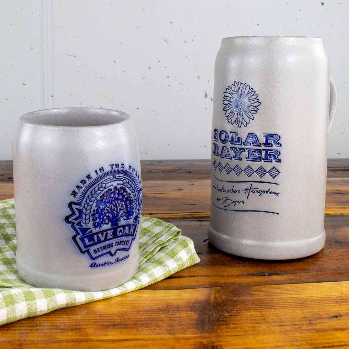 Zwei graue Steingut-Bierkrüge mit blauen Schriftzügen und Logos, stehend auf einem Holztisch.