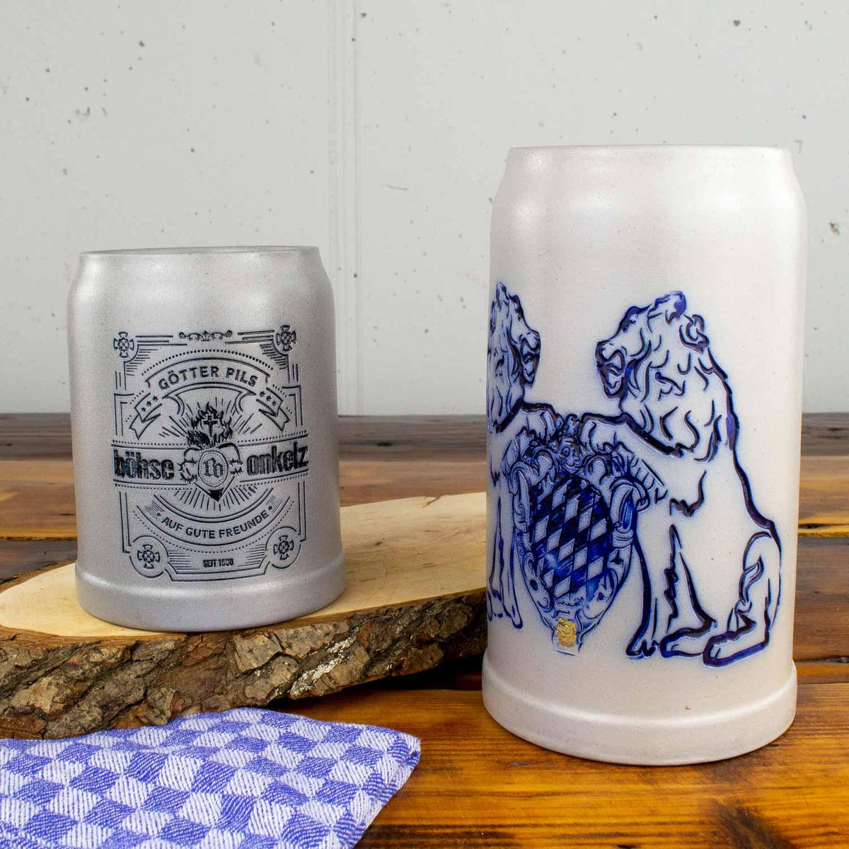 Zwei Keramik-Bierkrüge, ein heller mit blauem Löwenwappen und ein grauer mit dunklem Aufdruck auf Holzplatte, Küchenhandtuch.