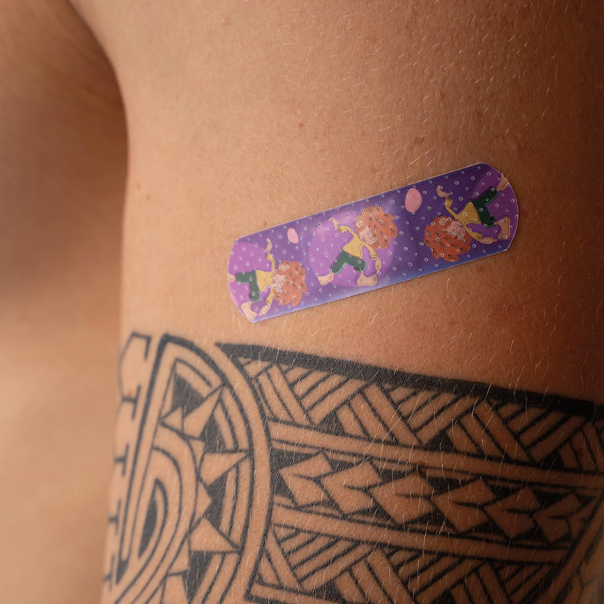 ®Pumuckl Trost-Pflaster auf behaarter Haut über einem Tattoo, lila mit Pumuckl-Motiven.
