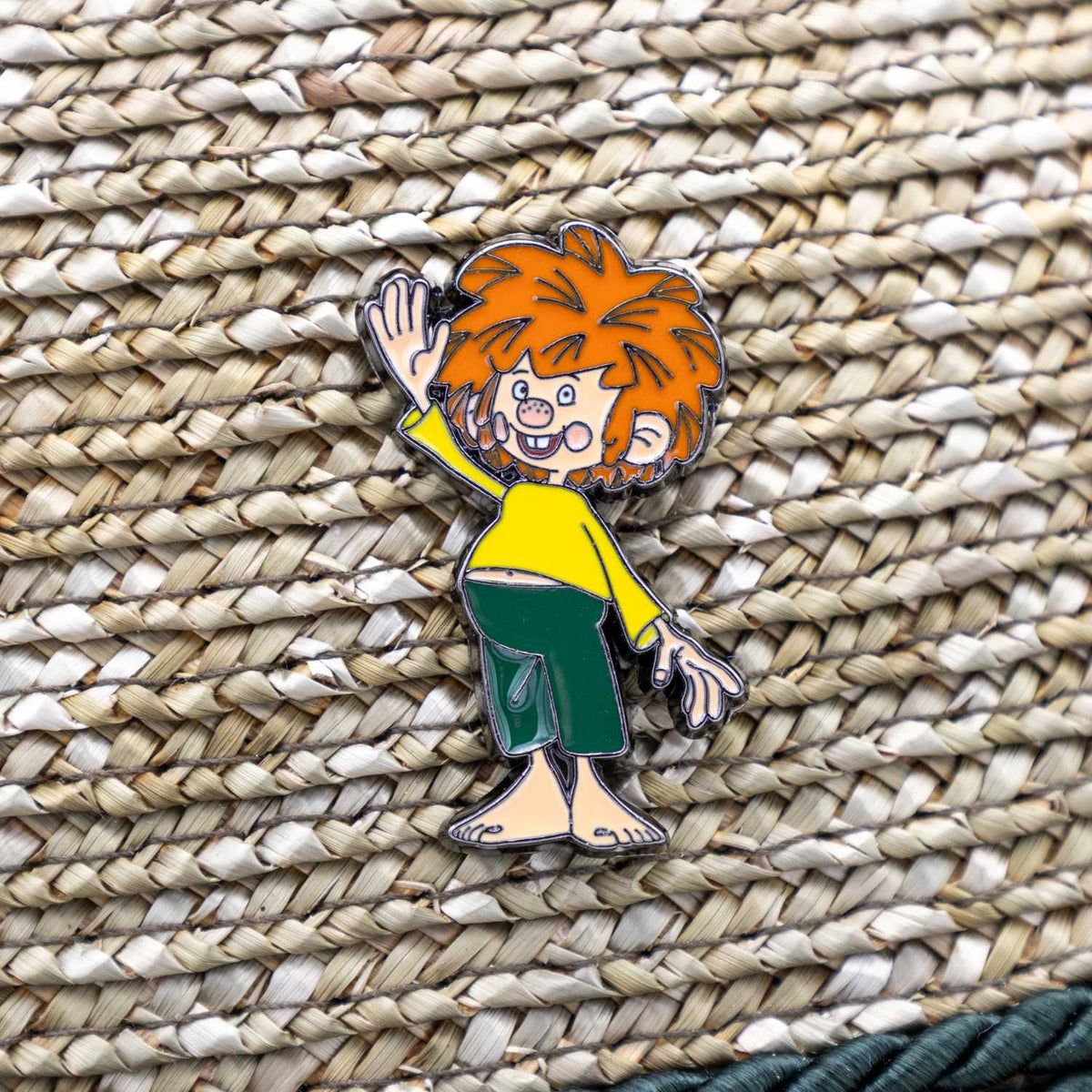 Pumuckl Servus Pin mit winkender Hand, auf hellem Korbgeflecht.