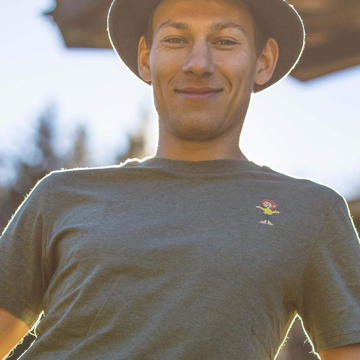 Lächelnder junger Mann mit Hut, trägt graues Pumuckl T-Shirt Pixmuckl als Brustprint in der Sonne.