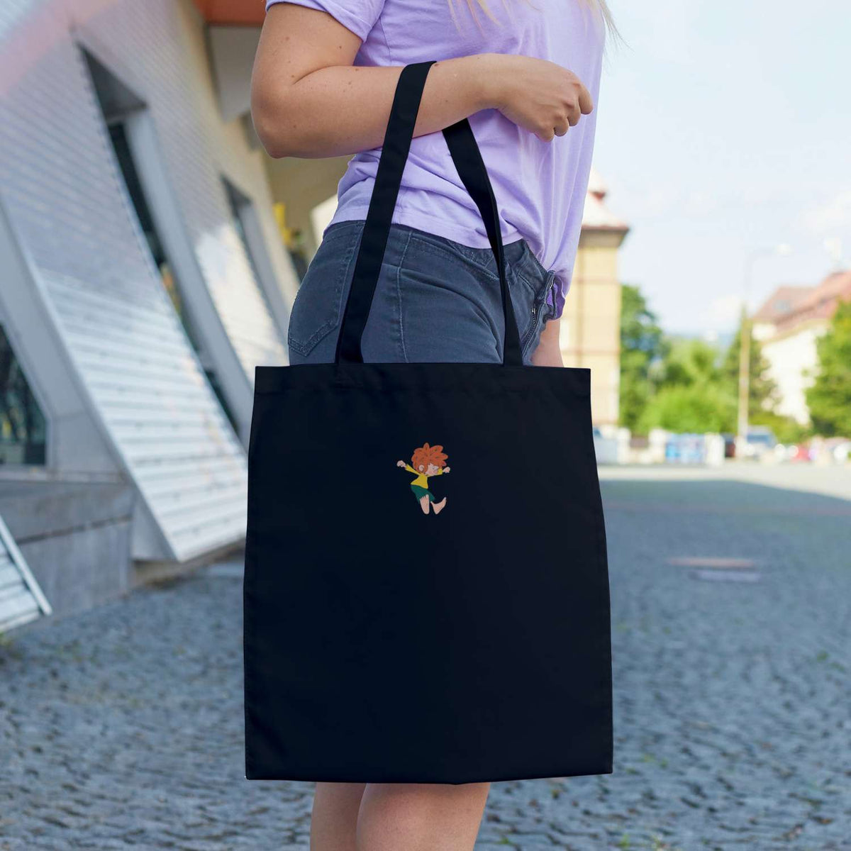 Frau trägt schwarze Pumuckl Tasche Bin a dabei mit lustigem Kobold-Print und violettem Shirt auf Kopfsteinpflaster.