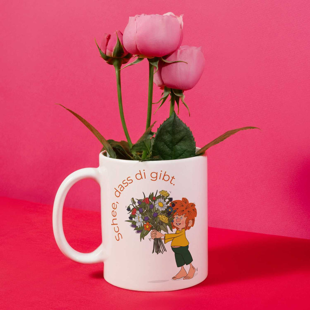 Weiße Pumuckl Tasse Schee,dass di gibt. mit frischen rosa Rosen vor pinkem Hintergrund.