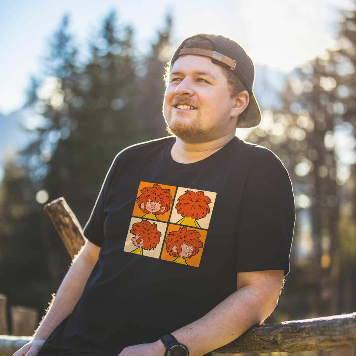 Lächelnder Mann mit Bart und Cap trägt schwarzes Pumuckl T-Shirt Schnappschuss in der Natur.