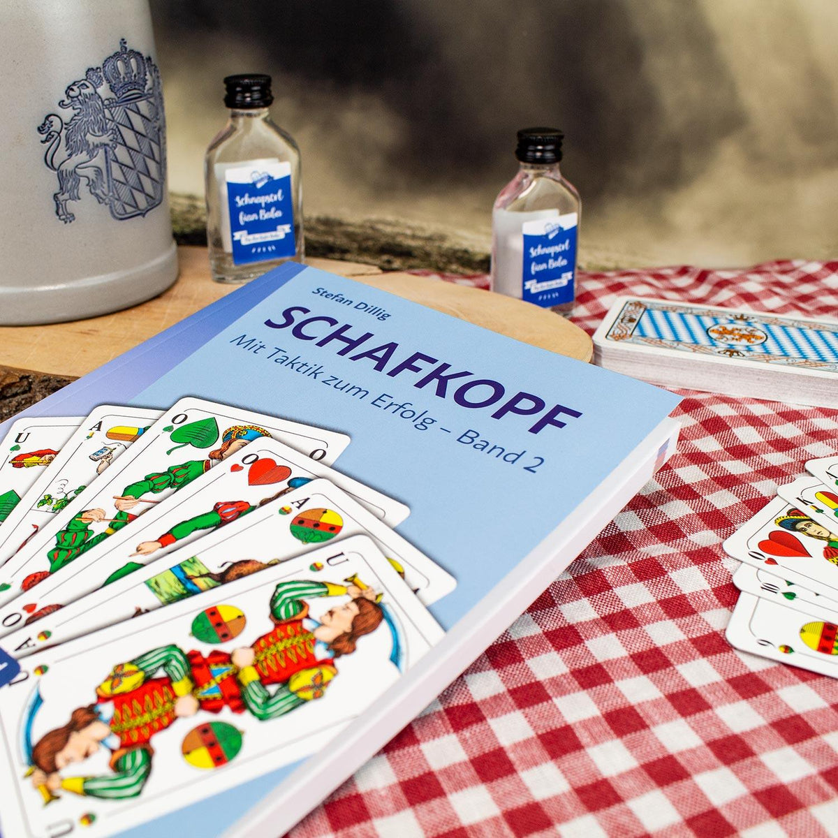 Schafkopf: Mit Taktik zum Erfolg – Band 2 Buch, umgeben von Spielkarten auf rot-weiß karierter Tischdecke.