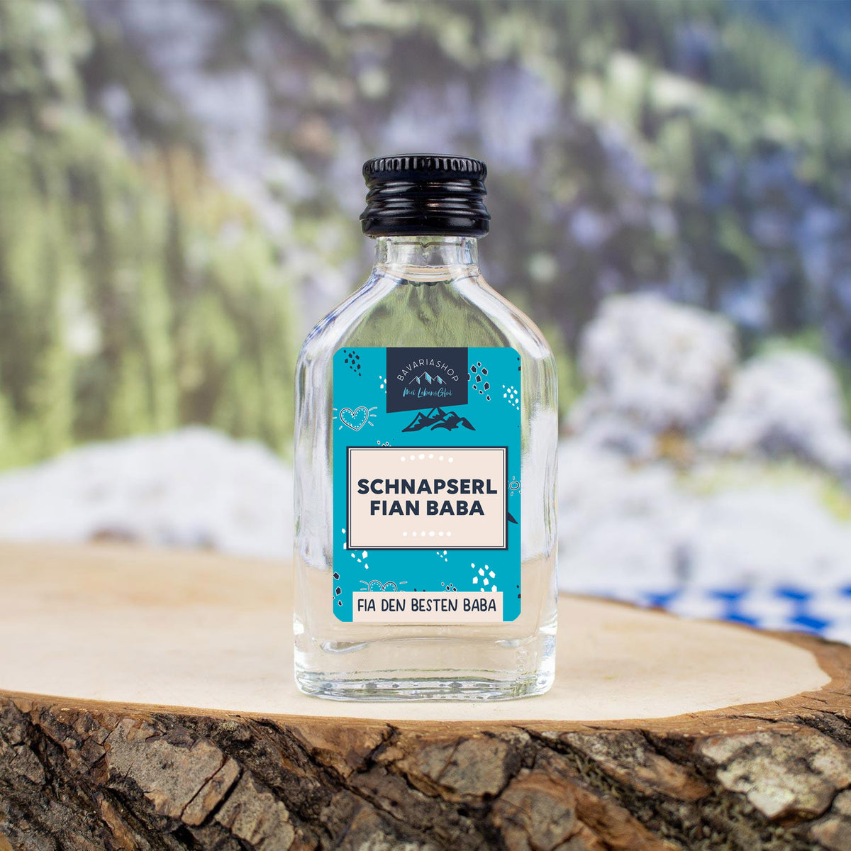 Klare Glasflasche Schnapserl Fian Baba mit blauem Etikett und schwarzem Drehverschluss auf einem Holzstück.