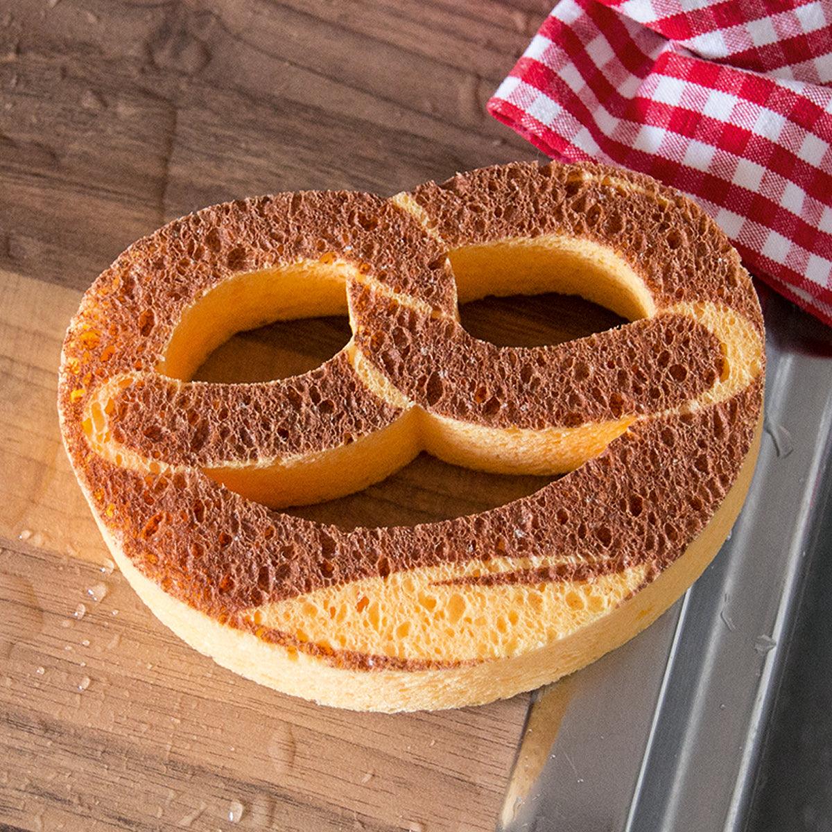Beiger Küchenschwamm in Form einer Brezel, auf Holztisch neben Spüle und rotem Geschirrtuch.