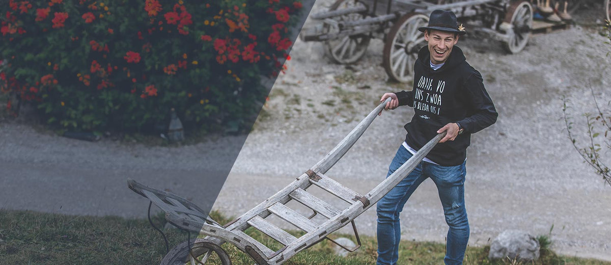 Fröhlicher junger Mann in schwarzem Kapuzenpullover und Hut zieht einen alten Leiterwagen, Kollektion für Buama.