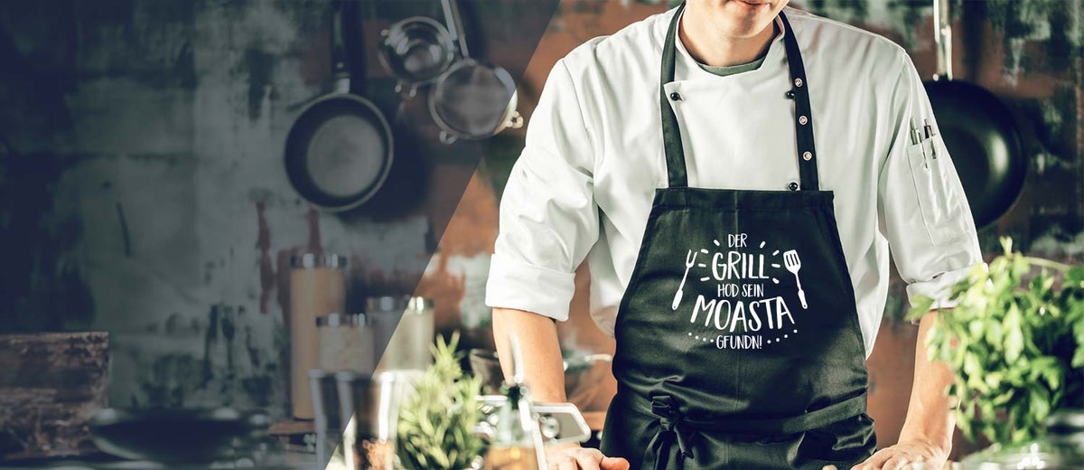 Mann in weißer Kochjacke und schwarzer Grillschürze mit bayerischem Spruch, umgeben von Küchenutensilien und Kräutern.