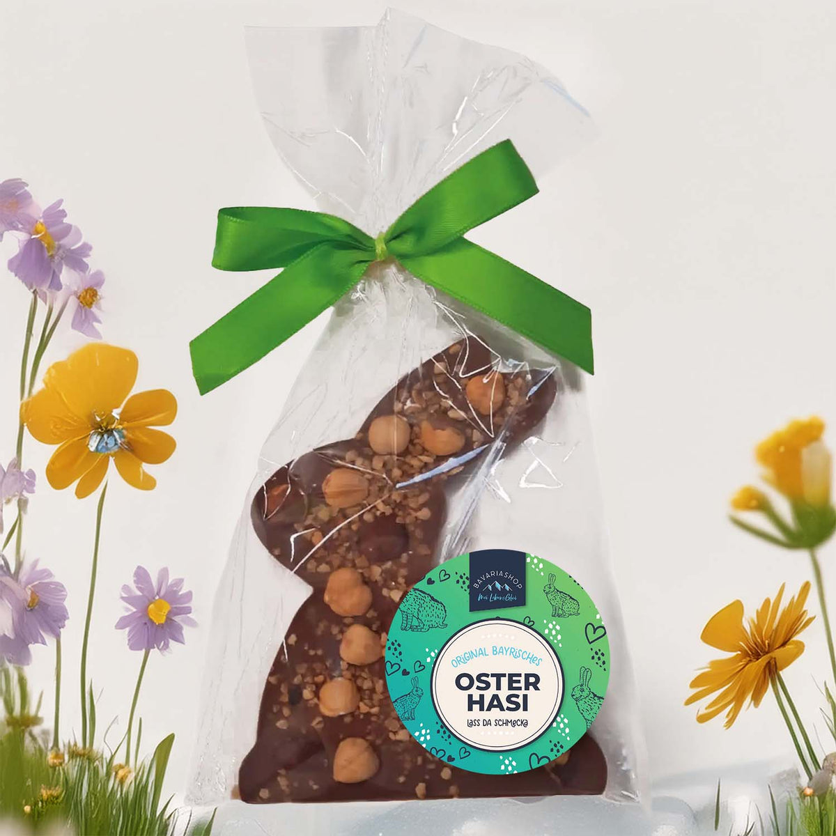 Oster-Schokoladen Nuss-Haserl in Klarsichtverpackung mit grüner Schleife, umgeben von bunten Frühlingsblumen.