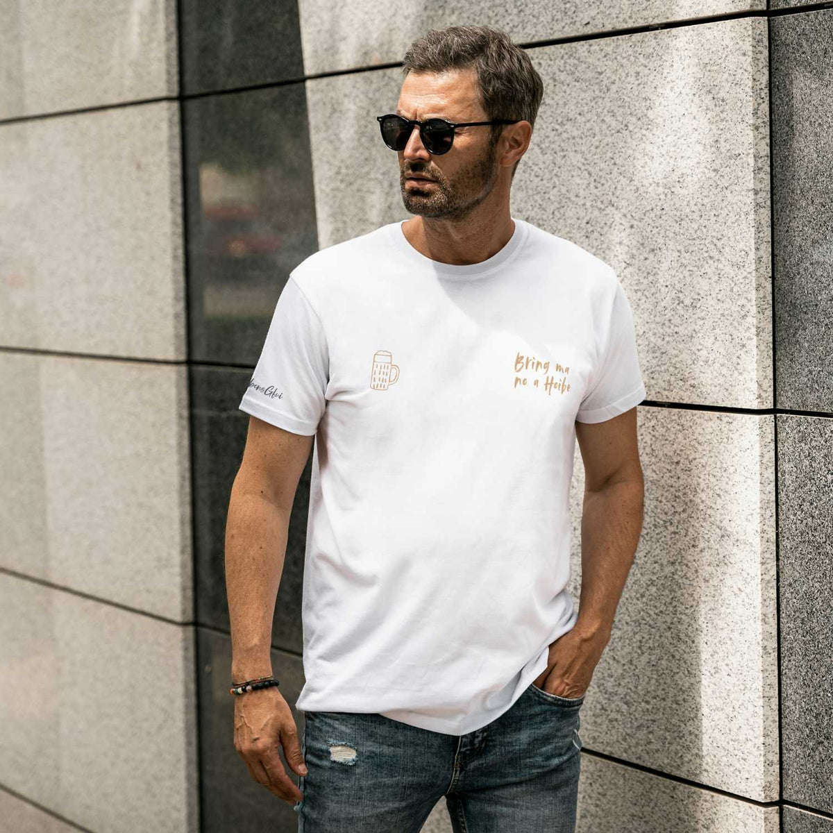 Herren T-Shirt Bring ma no a Hoibe in Weiß, goldener Druck und Bierkruglogo, urbaner Hintergrund.