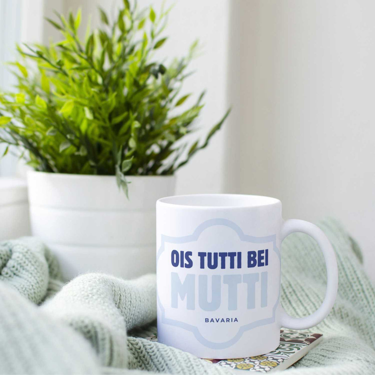 Weißer Kaffee Haferl Ois tutti bei Mutti mit bayerischem Spruch, auf Strickdecke vor Grünpflanze.