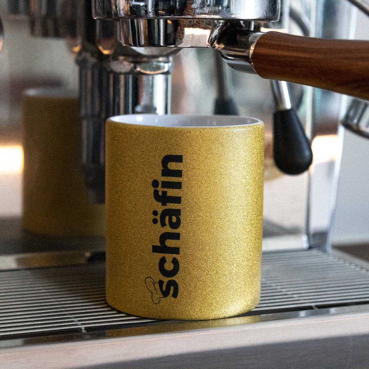 Kaffee Haferl Schäfin, gold glitzernde Tasse mit schwarzem Schriftzug unter einer Espressomaschine.