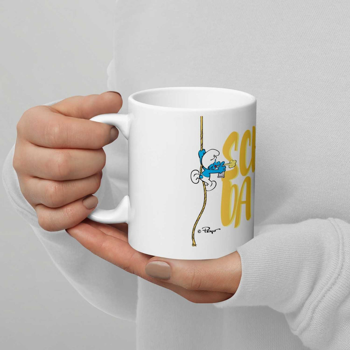 Weiße ®Die Schlümpfe Tasse Scheiß da nix mit Kletter-Schlumpf und gelbem Schriftzug in weiblichen Händen gehalten.